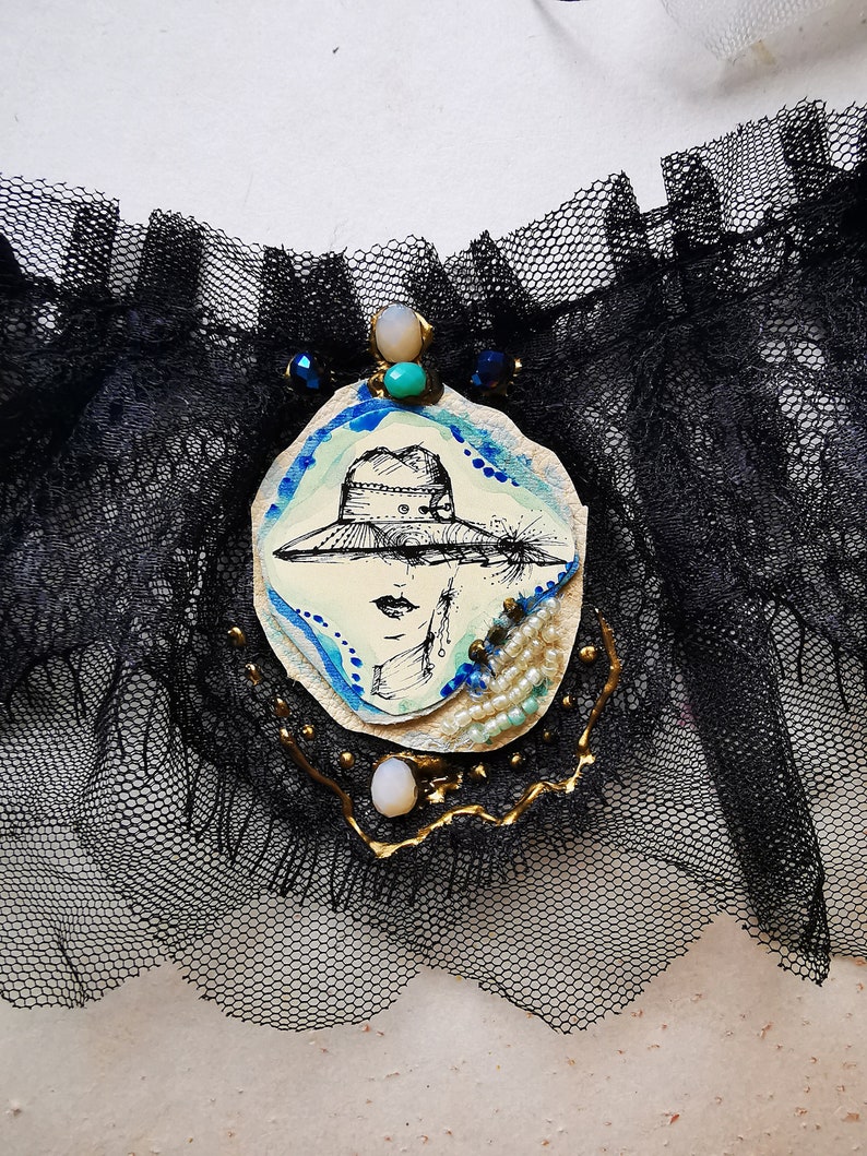 K&ouml;nnte beinhalten: Ein schwarzes Spitzenstrumpfband mit einem wei&szlig;en ovalen Kamee, das eine Frau mit Hut zeigt. Das Kamee ist mit blauen und t&uuml;rkisfarbenen Perlen und goldenen Akzenten verziert.