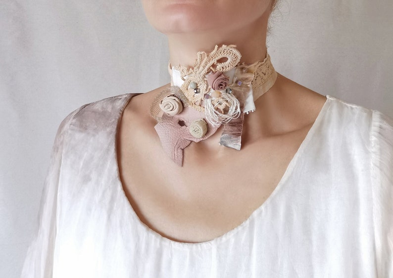 Peut inclure: Un d&eacute;licat collier ras de cou en dentelle blanche avec un grand ornement superpos&eacute; en tissu beige et rose, dentelle et perles. L'ornement pr&eacute;sente un motif floral avec une esth&eacute;tique vintage.