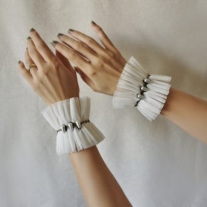 May include: Two white lace wrist cuffs with silver embellishments. The cuffs are made of delicate, ruffled fabric and feature a row of silver beads.