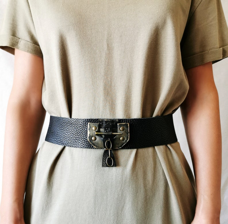 May include: A black leather belt with a large, ornate brass buckle featuring a keyhole design. The belt is worn over a light brown t-shirt.