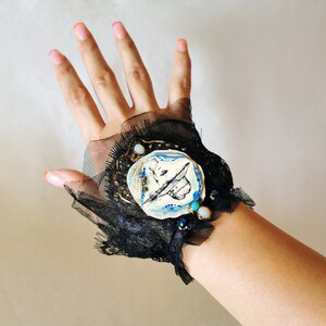 K&ouml;nnte beinhalten: Ein schwarzes Spitzen-Armband mit einem blauen und wei&szlig;en Keramik-Schmetterlings-Anh&auml;nger. Der Anh&auml;nger ist mit goldenen Akzenten und t&uuml;rkisfarbenen Perlen verziert.