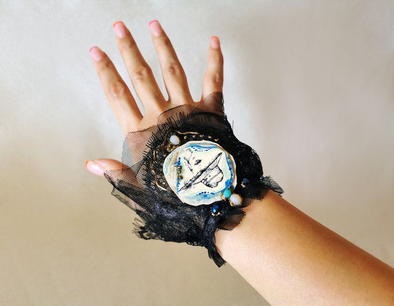 K&ouml;nnte beinhalten: Ein schwarzes Spitzen-Armband mit einem blauen und wei&szlig;en Keramik-Schmetterlings-Anh&auml;nger. Der Anh&auml;nger ist mit goldenen Akzenten und t&uuml;rkisfarbenen Perlen verziert.