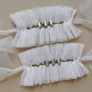 May include: Two white tulle wrist cuffs with gold trim and pearl accents. The cuffs are tied with white tulle ribbons.