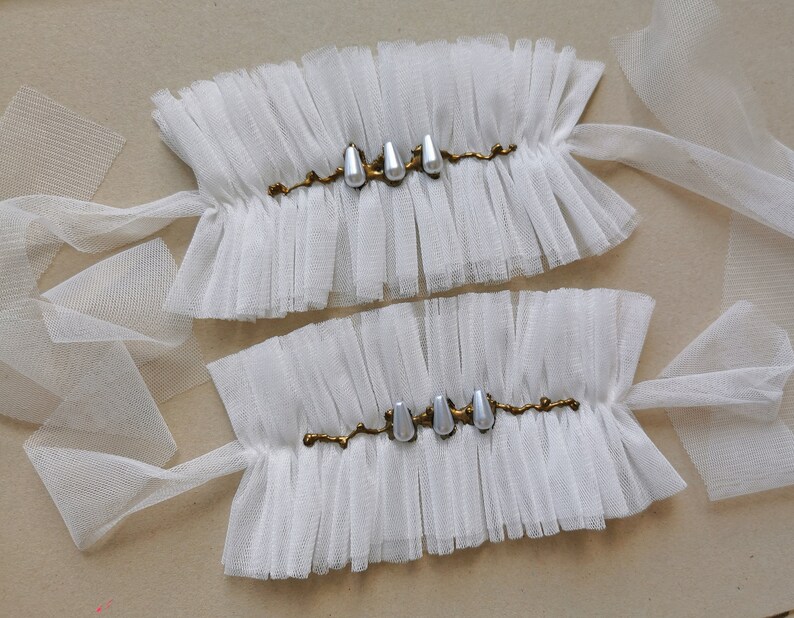 May include: Two white tulle wrist cuffs with gold trim and pearl accents. The cuffs are tied with white tulle ribbons.