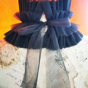 May include: A black tulle collar with a large bow in the front. The collar is made of multiple layers of tulle and has a ruffled edge.
