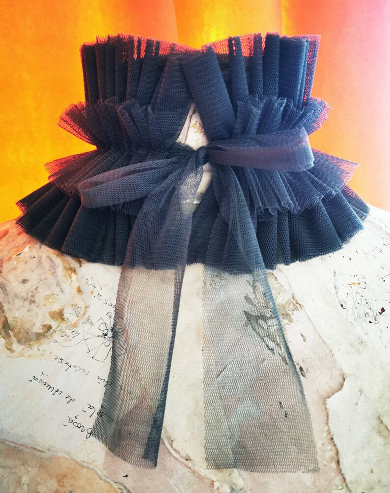 May include: A black tulle collar with a large bow in the front. The collar is made of multiple layers of tulle and has a ruffled edge.