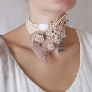 Peut inclure: Un collier ras de cou d&eacute;licat, fait main, compos&eacute; de diff&eacute;rents tissus, dont de la dentelle, du cuir et du coton. Le collier pr&eacute;sente un motif floral dans une palette de couleurs brun clair et blanc.