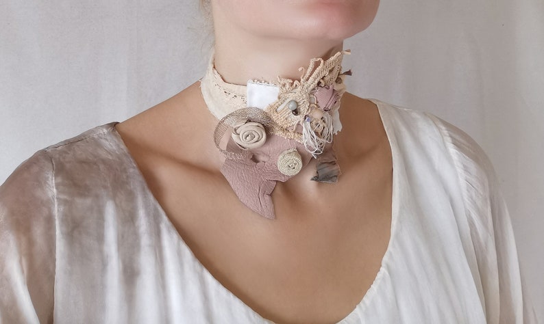 Peut inclure: Un collier ras de cou d&eacute;licat, fait main, compos&eacute; de diff&eacute;rents tissus, dont de la dentelle, du cuir et du coton. Le collier pr&eacute;sente un motif floral dans une palette de couleurs brun clair et blanc.