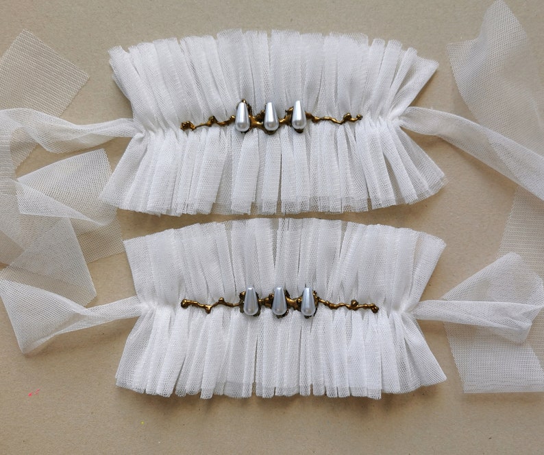 May include: Two white tulle wrist cuffs with gold embellishments and pearl-like accents.