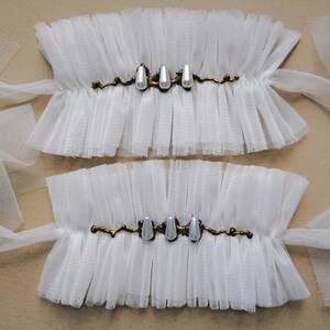 May include: Two white tulle wrist cuffs with gold trim and pearl accents. The cuffs are tied with white ribbon.