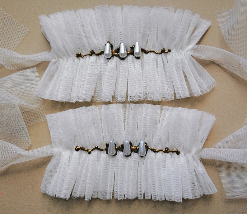 May include: Two white tulle wrist cuffs with gold trim and pearl accents. The cuffs are tied with white ribbon.