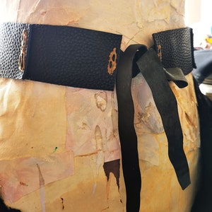 May include: A black leather belt with a decorative metal buckle and a long black ribbon tie. The belt is tied around a mannequin.