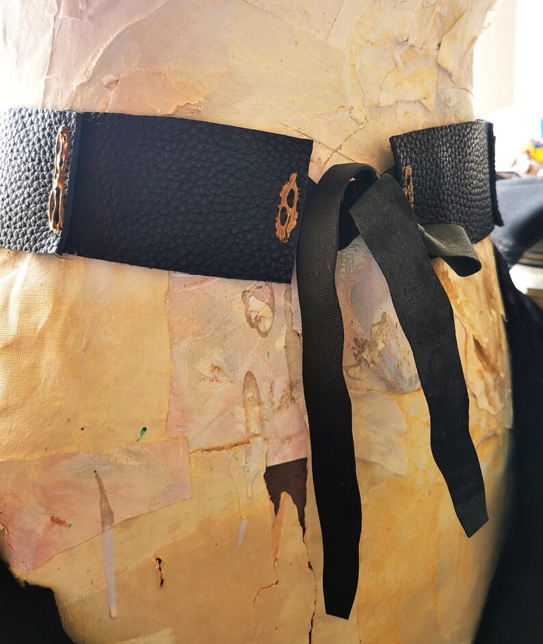 May include: A black leather belt with a decorative metal buckle and a long black ribbon tie. The belt is tied around a mannequin.