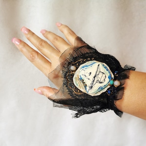 K&ouml;nnte beinhalten: Ein schwarzes Spitzen-Armband mit einem wei&szlig;en Keramik-Anh&auml;nger, der eine handgezeichnete Illustration eines Hutes zeigt. Der Anh&auml;nger ist mit goldenen und blauen Akzenten und kleinen Perlen verziert.