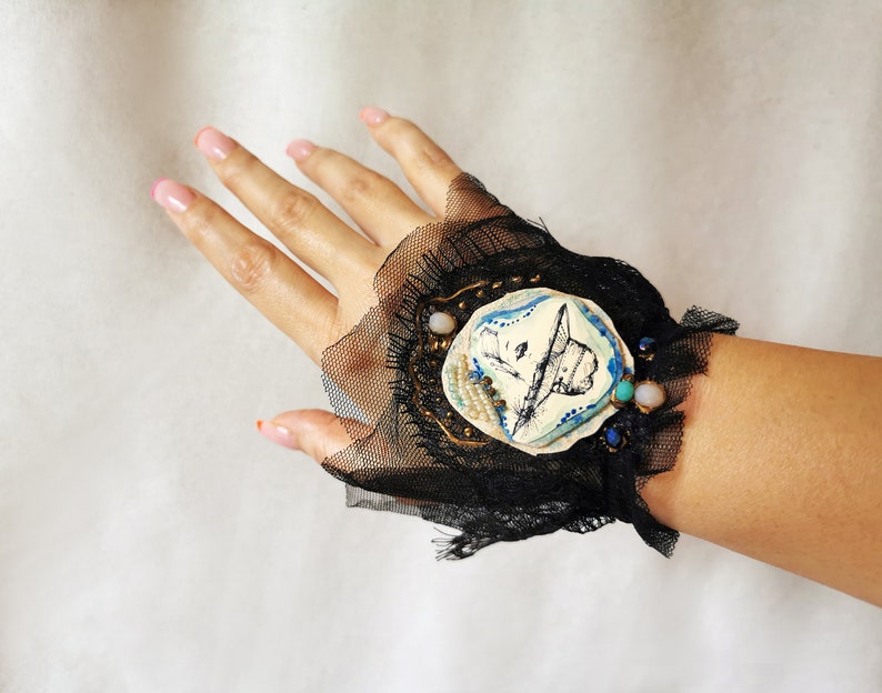 K&ouml;nnte beinhalten: Ein schwarzes Spitzen-Armband mit einem wei&szlig;en Keramik-Anh&auml;nger, der eine handgezeichnete Illustration eines Hutes zeigt. Der Anh&auml;nger ist mit goldenen und blauen Akzenten und kleinen Perlen verziert.