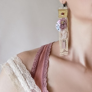 Può includere: Orecchini pendenti lunghi con un design unico e misto. Gli orecchini presentano una combinazione di tessuto dorato, elementi strutturati e accenti decorativi nei toni del rosa, lavanda e crema. Gli orecchini sono attaccati a un gancio color argento.