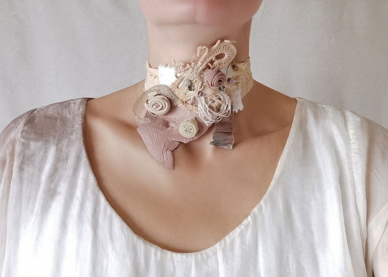 Peut inclure: Un collier ras de cou d&eacute;licat et superpos&eacute; en tissu, dentelle et cuir. Le collier pr&eacute;sente une vari&eacute;t&eacute; de textures et de couleurs, notamment le blanc, le beige et le brun. Le collier est orn&eacute; de petites fleurs d&eacute;licates et de perles.