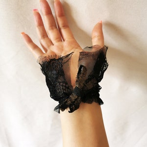 K&ouml;nnte beinhalten: Schwarze Spitzen-Handgelenksmanschetten mit einem goldenen Schleifendetail. Die Manschetten sind aus zarter Spitze und T&uuml;ll gefertigt.