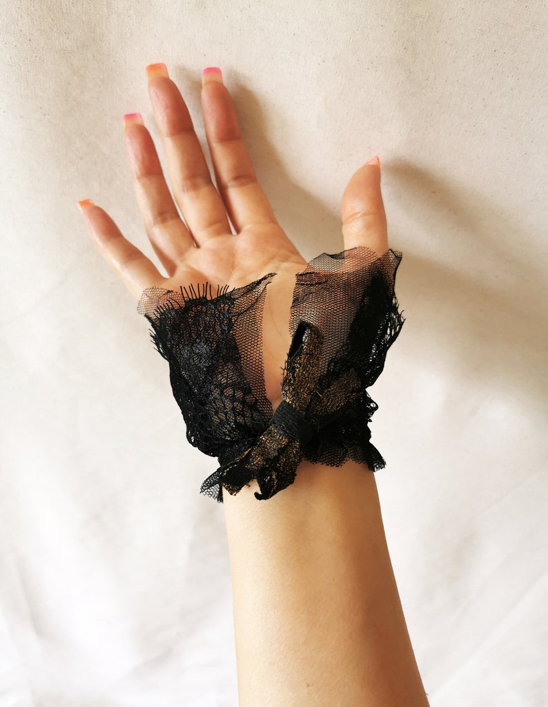 K&ouml;nnte beinhalten: Schwarze Spitzen-Handgelenksmanschetten mit einem goldenen Schleifendetail. Die Manschetten sind aus zarter Spitze und T&uuml;ll gefertigt.