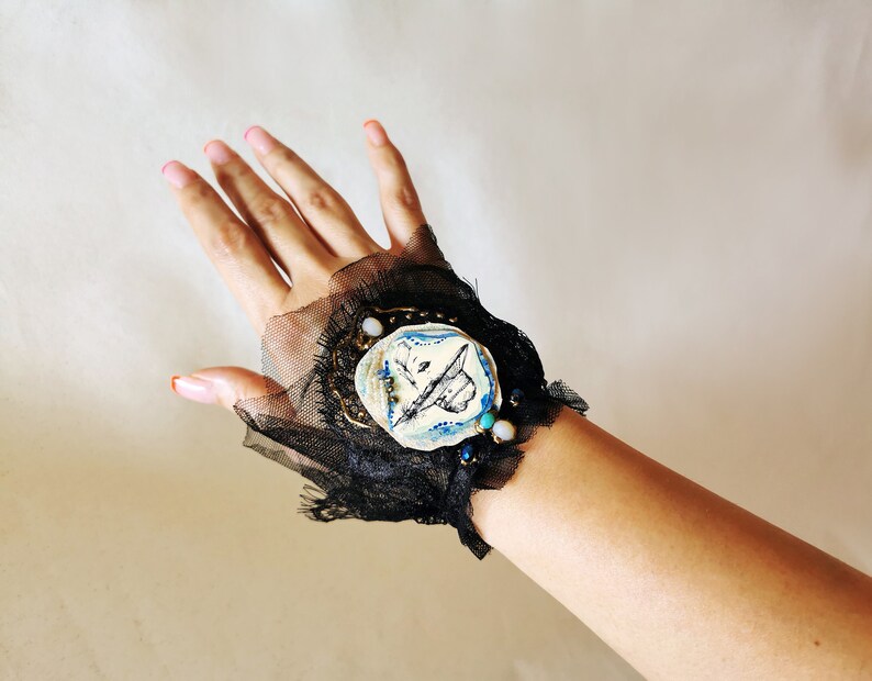 K&ouml;nnte beinhalten: Ein schwarzes Spitzen-Armband mit einem wei&szlig;en Keramik-Anh&auml;nger mit einem blauen und schwarzen Design. Der Anh&auml;nger ist mit goldenen und blauen Perlen verziert.