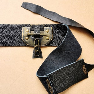 May include: A black leather belt with a brass buckle and a decorative chain. The buckle has a screw-like design and is attached to the belt with two rivets.