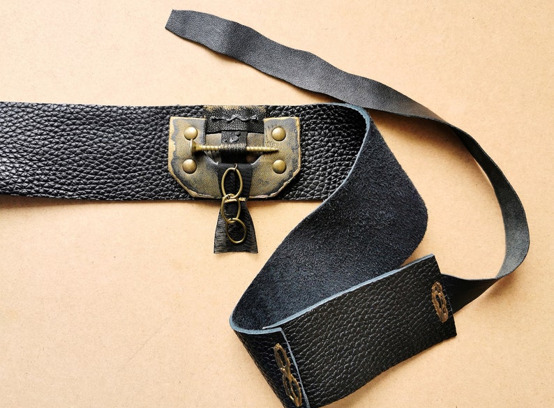 May include: A black leather belt with a brass buckle and a decorative chain. The buckle has a screw-like design and is attached to the belt with two rivets.