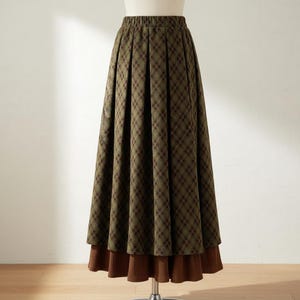 May include: A long, pleated skirt featuring an olive green and dark brown plaid pattern. The skirt has an elasticized waistband and a solid brown, ribbed-texture hem panel that extends below the main plaid fabric, creating a layered appearance.