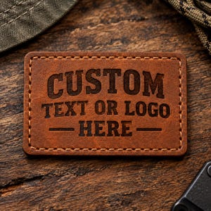 May include: A rectangular brown leather patch with rounded corners and visible stitching along the edges. The patch features the laser-engraved text "CUSTOM TEXT OR LOGO HERE" in a bold, dark, sans-serif font centred on the surface.