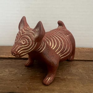 May include: A terracotta-colored ceramic dog figurine featuring carved, light-colored swirling and linear patterns across its body, head, and ears. The sculpture depicts a four-legged animal in a standing position with pointed ears and a short tail.