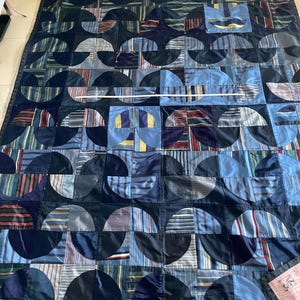 May include: A quilt featuring a repeating pattern of dark blue semicircles and rectangular blocks made from various striped and solid blue fabrics. A white measuring tape is stretched horizontally across the centre of the quilt.
