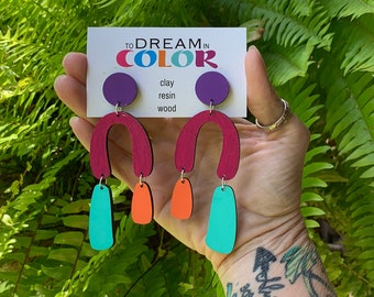 May include: A pair of colorful earrings with a geometric design. The earrings are made of clay, resin, and wood. The earrings are in shades of red, orange, turquoise, and purple. The earrings are being held by a hand with a tattoo.