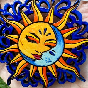 May include: A pair of wooden earrings with a blue, yellow, and orange sun and moon design. The sun and moon are painted with faces and have a blue and white background.
