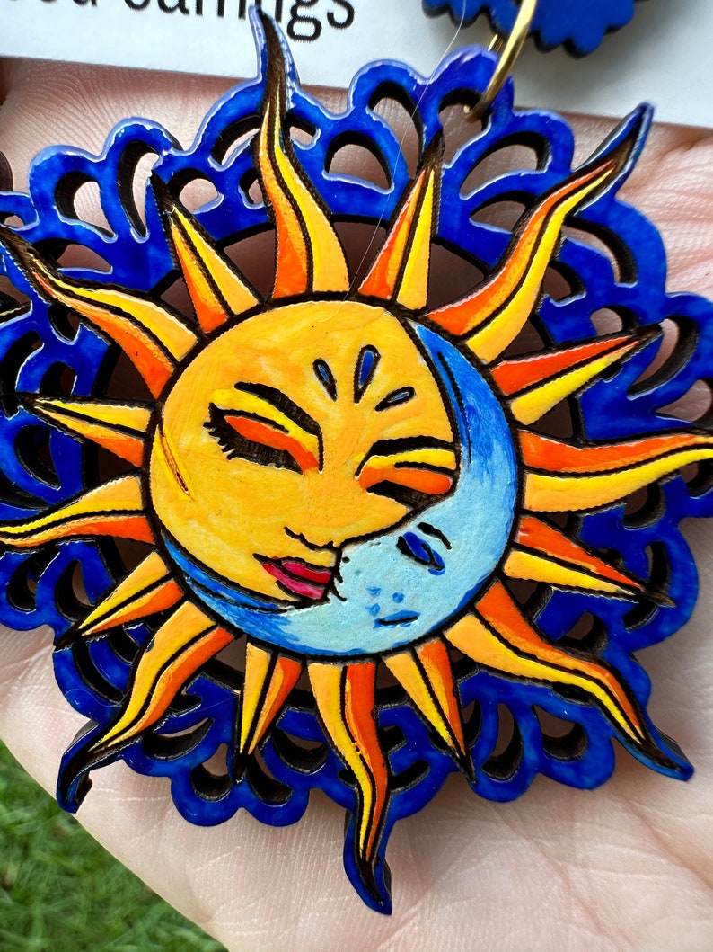 May include: A pair of wooden earrings with a blue, yellow, and orange sun and moon design. The sun and moon are painted with faces and have a blue and white background.