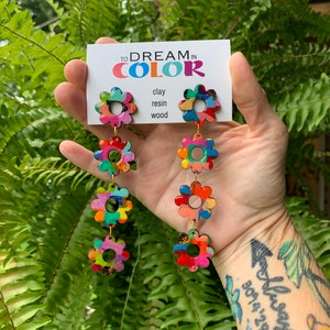 May include: A pair of colorful flower-shaped earrings with a layered design. The earrings are made of clay, resin, and wood. The text on the card reads "To Dream in Color".