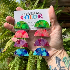 May include: A pair of colorful, multi-layered, wooden earrings with gold accents. The earrings are painted in shades of green, pink, purple, and blue. The earrings are displayed against a background of green ferns. The card in the image reads "TO DREAM IN COLOR" and "One of a kind and painted wood earrings".