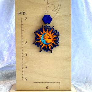 May include: A wooden sun and moon earring with a blue and orange color scheme. The earring is about 2 inches long and features a detailed design of a sun and moon with a face.