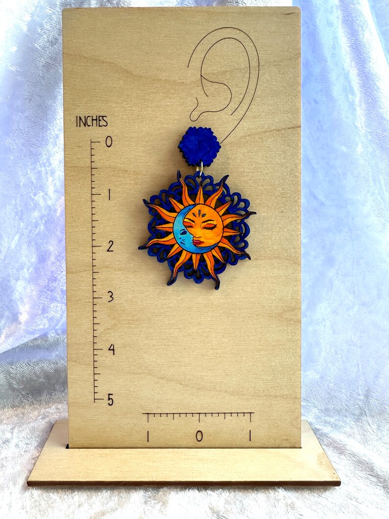May include: A wooden sun and moon earring with a blue and orange color scheme. The earring is about 2 inches long and features a detailed design of a sun and moon with a face.