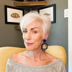 May include: A woman with short, white hair wearing a gray off-the-shoulder shirt and large, blue and gold sun and moon earrings.