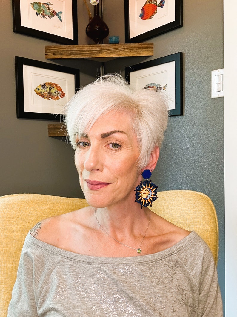 May include: A woman with short, white hair wearing a gray off-the-shoulder shirt and large, blue and gold sun and moon earrings.