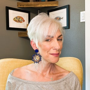 May include: A woman with short, silver hair wearing a gray shirt and large, blue and gold sun earrings. The earrings feature a sun with a face and a moon with a face.