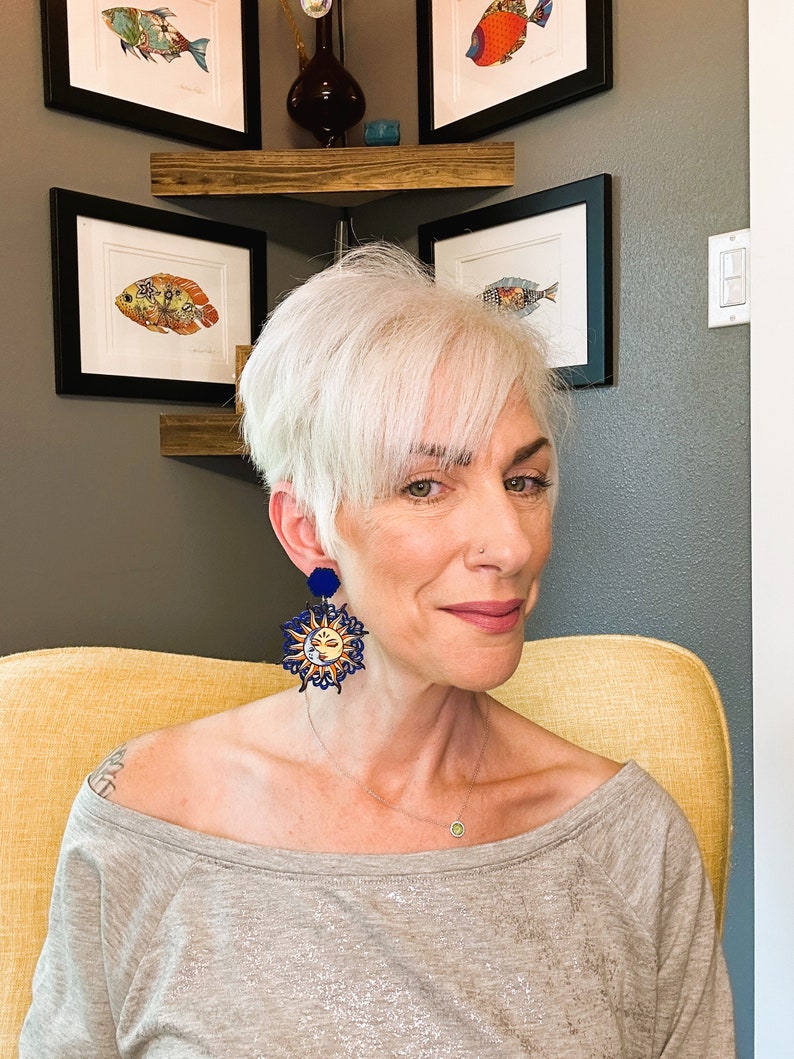 May include: A woman with short, silver hair wearing a gray shirt and large, blue and gold sun earrings. The earrings feature a sun with a face and a moon with a face.