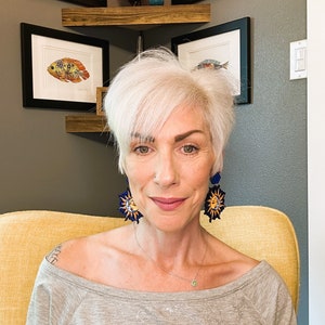 May include: A woman with short, white hair is wearing a gray off-the-shoulder shirt and a pair of earrings with a sun and moon design. The earrings are blue and yellow.