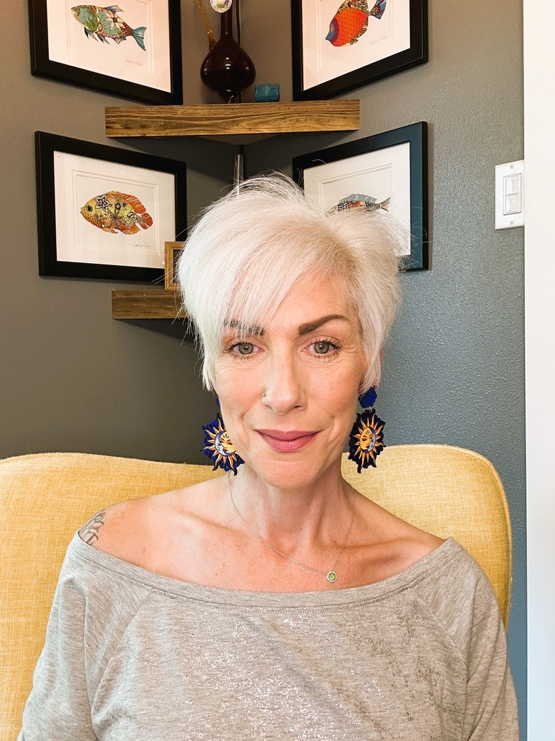 May include: A woman with short, white hair is wearing a gray off-the-shoulder shirt and a pair of earrings with a sun and moon design. The earrings are blue and yellow.