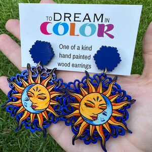 May include: A pair of hand-painted wooden earrings featuring a sun and moon design. The earrings are blue, yellow, and orange. The earrings are labeled "To Dream in Color" and "One of a kind hand painted wood earrings".