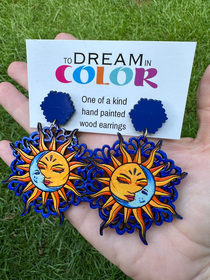 May include: A pair of hand-painted wooden earrings featuring a sun and moon design. The earrings are blue, yellow, and orange. The earrings are labeled "To Dream in Color" and "One of a kind hand painted wood earrings".