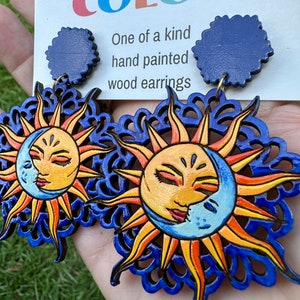 May include: A pair of hand-painted wooden earrings featuring a sun and moon design. The earrings are blue, yellow, orange, and white. The earrings are attached to blue circular posts. The card behind the earrings reads "To Dream in Color" and "One of a kind hand painted wood earrings".