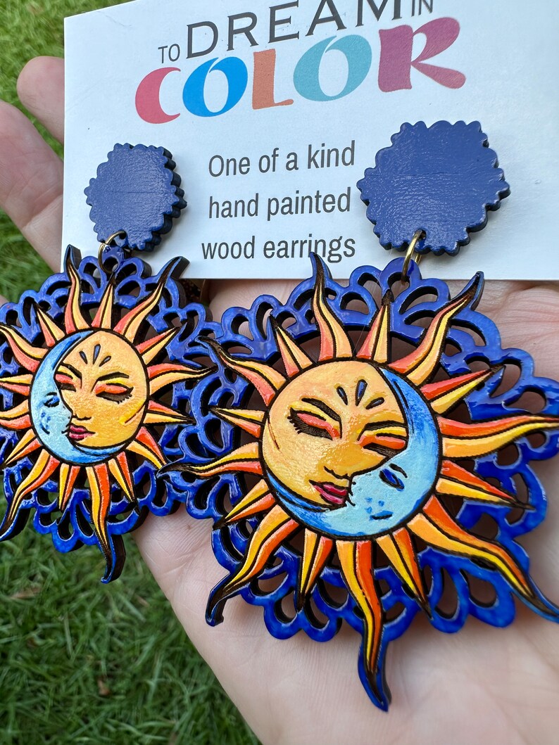 May include: A pair of hand-painted wooden earrings featuring a sun and moon design. The earrings are blue, yellow, orange, and white. The earrings are attached to blue circular posts. The card behind the earrings reads "To Dream in Color" and "One of a kind hand painted wood earrings".