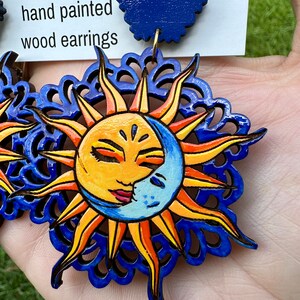 May include: A pair of hand-painted wooden earrings featuring a sun and moon design. The sun is yellow with a smiling face and the moon is blue with a crescent shape. The earrings are surrounded by a blue and orange mandala design.