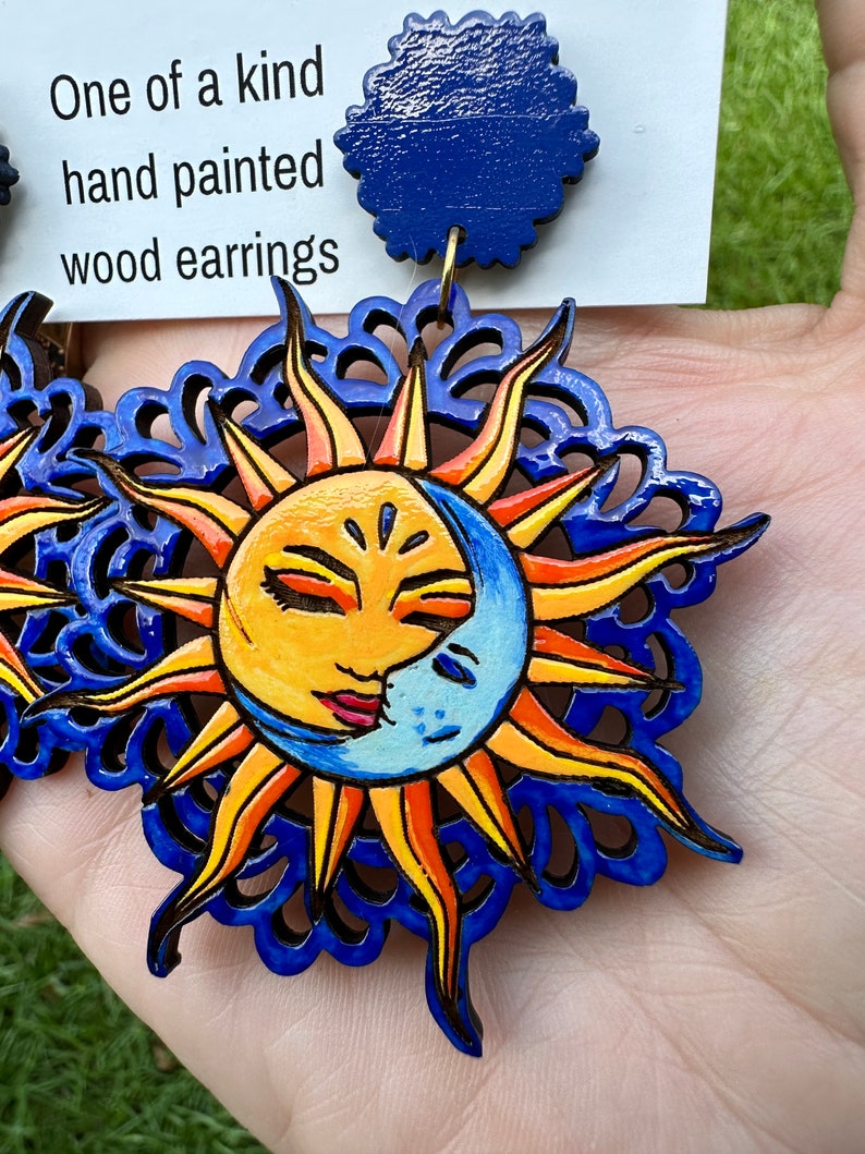 May include: A pair of hand-painted wooden earrings featuring a sun and moon design. The sun is yellow with a smiling face and the moon is blue with a crescent shape. The earrings are surrounded by a blue and orange mandala design.