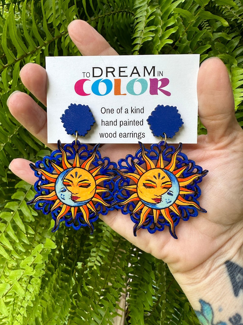 May include: A pair of hand-painted wooden earrings featuring a sun and moon design. The earrings are blue, yellow, and orange with a detailed design. The earrings are labeled "To Dream in Color" and "One of a kind hand painted wood earrings".